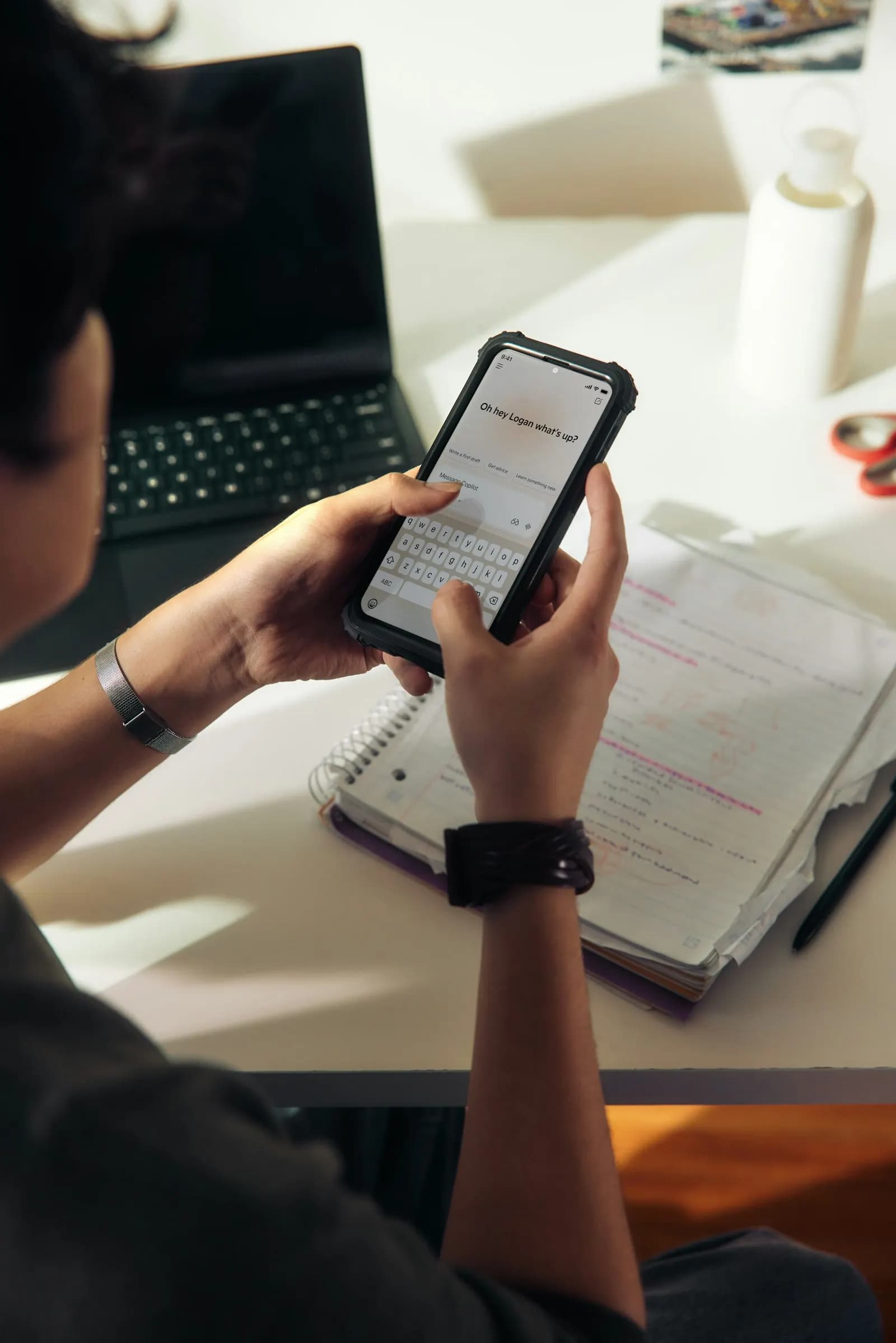 A person checks a learning app on their phone, sitting at a desk with an open notebook.